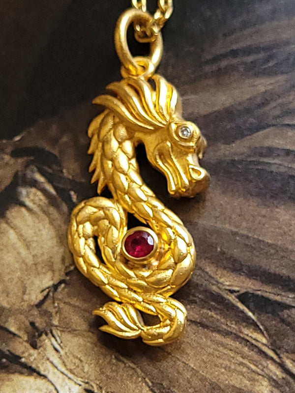 A large gold dragon pendant with a central bezel set ruby and two tiny diamonds in the eyes, on a separate chain.
