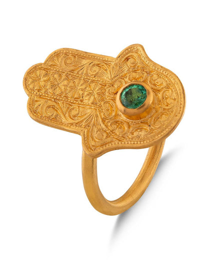 A gold-colored, palm-shaped Hamsa ring with a central emerald cabochon, surrounded by intricate leaf patterns.